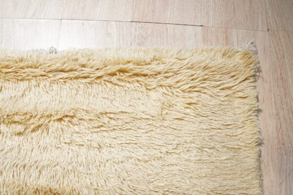 The edge of a pale yellow, long-pile shag rug rests on light blonde wood planks.