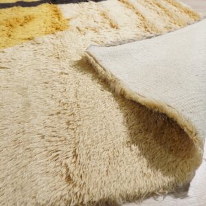 Shaggy rug in cream and yellow stripes, curled back to reveal its woven backing on wood floor.