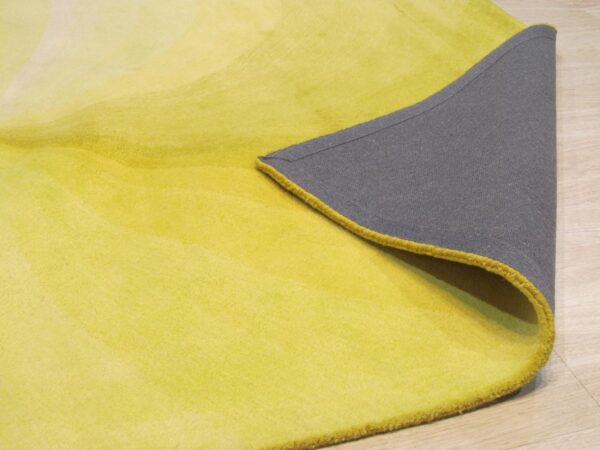 A yellow gradient rug pile is folded back, revealing a gray backing and bright yellow edge binding on light wood flooring.