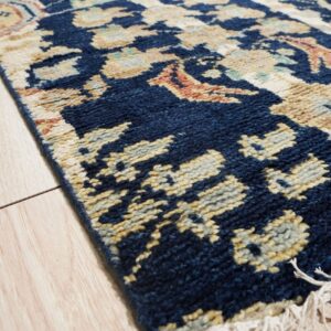 Textured navy blue rug with traditional cream and gold patterns and fringes on light wood plank flooring.