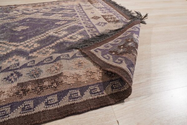 Purple and brown geometric rug with visible fringes, partially folded back on light wood planks.