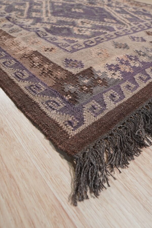 Textured rug with purple, brown, and beige geometric patterns, featuring fringe, lying on light wood flooring.