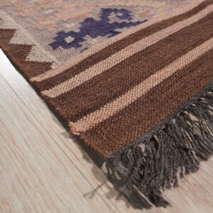 Flat woven rug corner showing purple motifs, brown stripes, and dark gray fringe on light wood flooring.