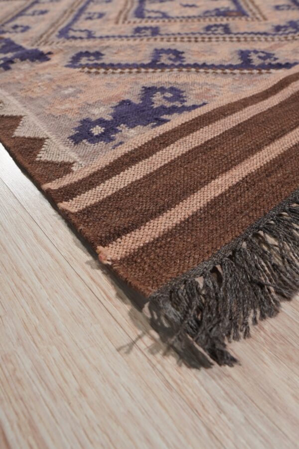 Flat woven rug corner showing purple motifs, brown stripes, and dark gray fringe on light wood flooring.