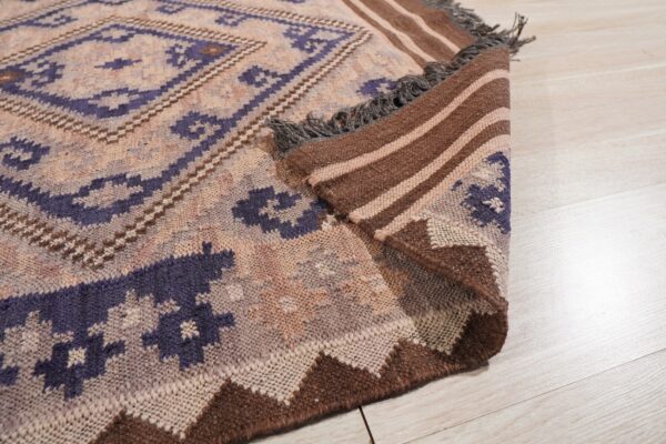 A purple and tan geometric rug is folded on light flooring, showing a striped brown backing and fringe.