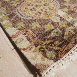 A chunky-pile rug with brown, cream, and gold traditional patterns and fringe rests on light plank flooring.
