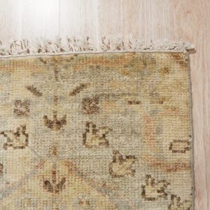 Corner of a textured beige rug with dark patterns and cream fringe on blonde wood flooring.