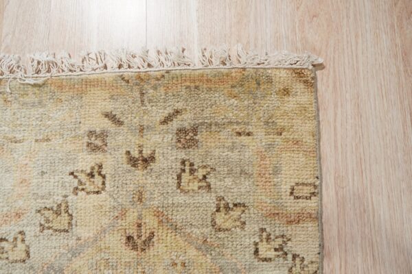 Corner of a textured beige rug with dark patterns and cream fringe on blonde wood flooring.