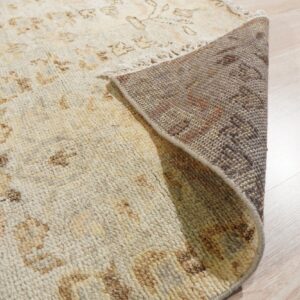 Light beige patterned rug corner rolled up, showing the dark woven backing and light wood flooring.
