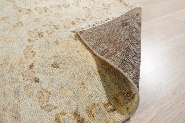 Light beige patterned rug corner rolled up, showing the dark woven backing and light wood flooring.