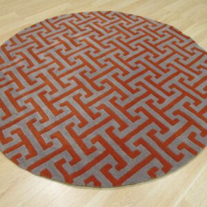 Round rug with a gray and terracotta geometric key pattern and visible texture on light wood flooring.