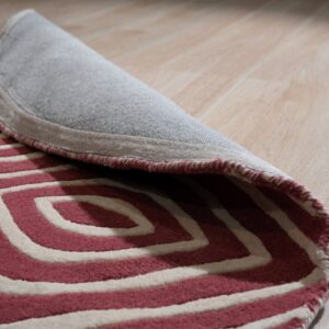 The edge of a pink and cream textured rug with concentric patterns is rolled up on wood plank flooring.