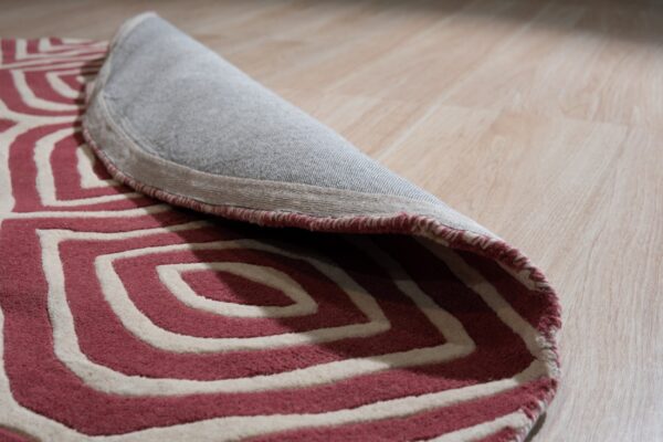 The edge of a pink and cream textured rug with concentric patterns is rolled up on wood plank flooring.
