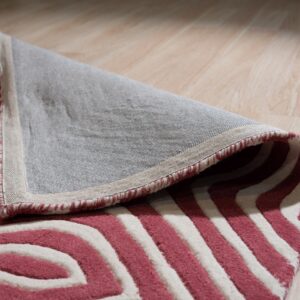 A pink and white high-pile geometric rug is folded back, revealing the woven backing on light wood flooring.