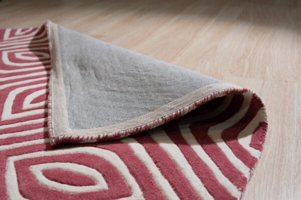 A pink and white high-pile geometric rug is folded back, revealing the woven backing on light wood flooring.