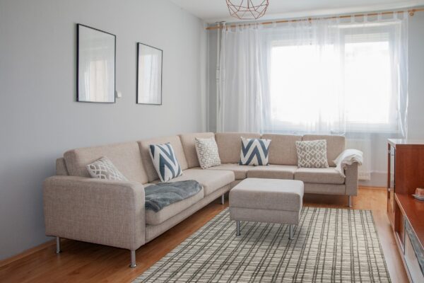 Gray and beige grid pattern rug partially under a large light-colored sectional sofa on hardwood floor.