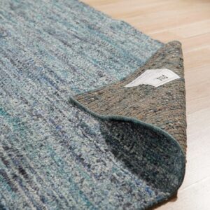 Highly textured blue and gray rug lifted to expose the brown woven backing lying on light wood flooring.