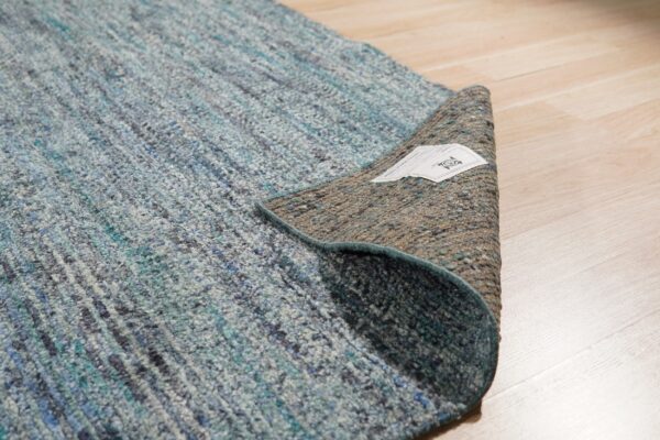 Highly textured blue and gray rug lifted to expose the brown woven backing lying on light wood flooring.