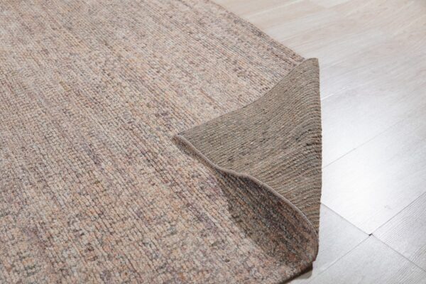 Low pile, mottled beige rug with the corner flipped to show the dark backing on a light wood floor.
