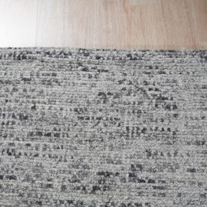 A highly textured gray and charcoal speckled rug lies on light natural wood plank flooring.