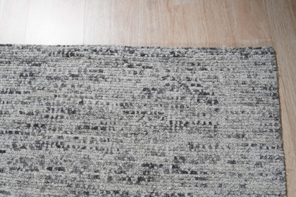 A highly textured gray and charcoal speckled rug lies on light natural wood plank flooring.