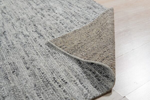 Speckled gray textured rug pile and woven tan backing displayed on light wood floorboards.