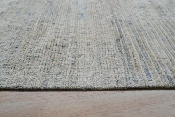 A textured, high-pile beige rug with blue speckles rests on light colored plank wood flooring.