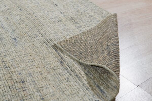 Textured light beige rug partially rolled back on light wood flooring, revealing its woven brown underside.