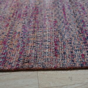 Close-up of a flecked rug in magenta, navy, and earth tones resting on light wood plank flooring.