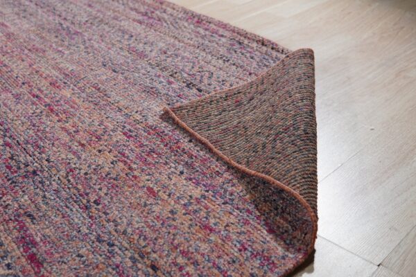 Flecked magenta, purple, and beige rug corner is lifted, revealing the dark woven backing.