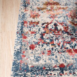 Low pile rug with an abstract design featuring cream, blue, red, and gold over light wood plank flooring.