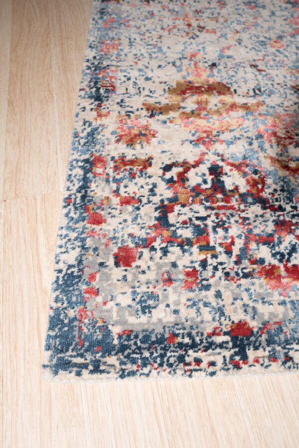Low pile rug with an abstract design featuring cream, blue, red, and gold over light wood plank flooring.