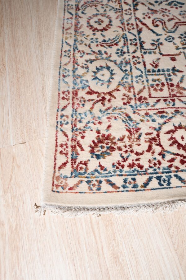 Cream rug with intricate red and blue traditional designs shown close-up on light blonde wood flooring.