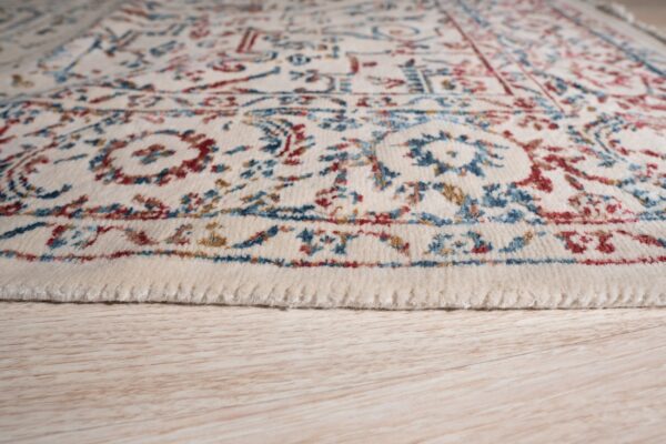 Medium pile cream rug with red and blue traditional motifs resting on light wood grain floor.
