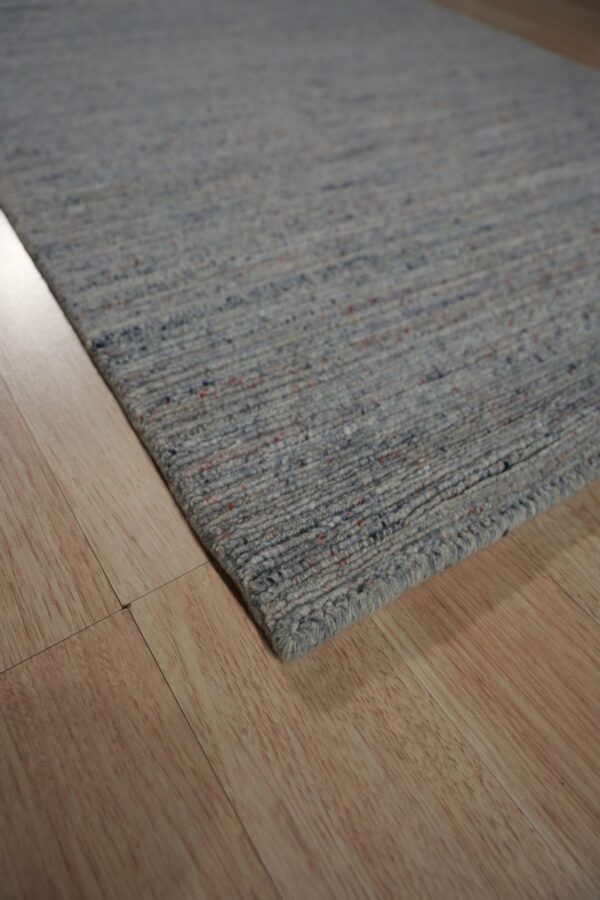 Speckled gray, low-pile rug with visible blue and red fibers rests on light wood plank flooring.