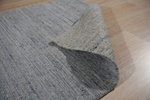 Close-up of a ribbed blue-gray rug, showing the colorful flecked pile and rolled backing on light wood flooring.