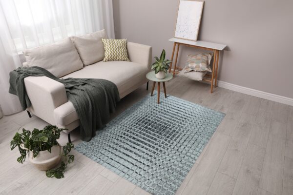 A light blue and dark grey grid-pattern rug lies under a beige sofa on light wood flooring.