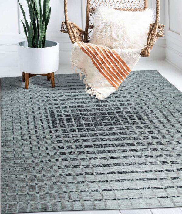 A gray and black distressed geometric rug rests on a white floor next to a rattan chair and snake plant.