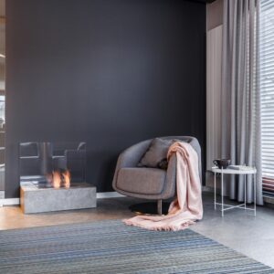 Striped blue and gray low-pile rug beneath a modern armchair, a pink throw, and a lit fireplace.
