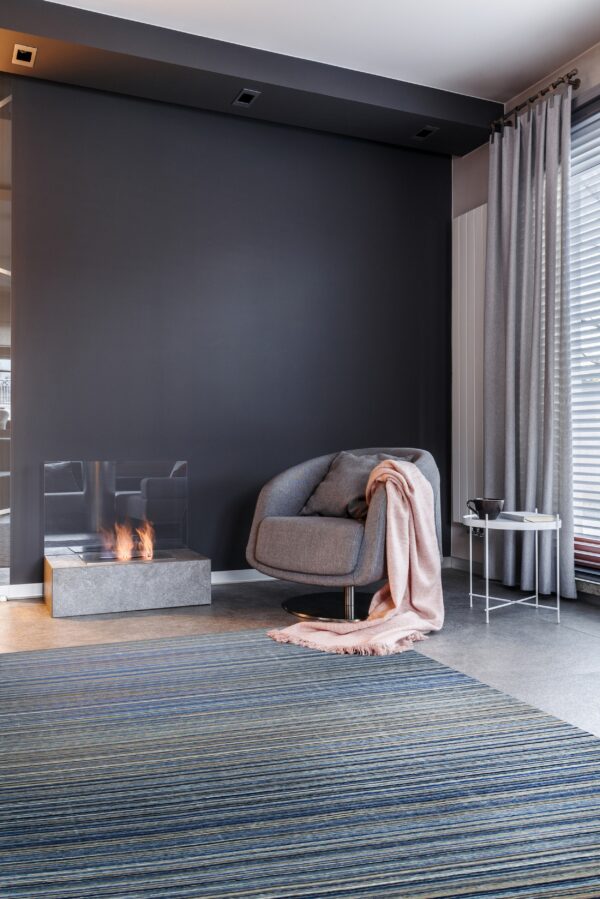 Striped blue and gray low-pile rug beneath a modern armchair, a pink throw, and a lit fireplace.