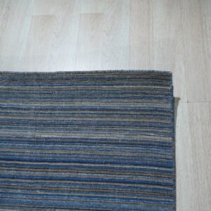 A corner view of a low-pile rug with blue, brown, and tan horizontal stripes on light wood flooring.