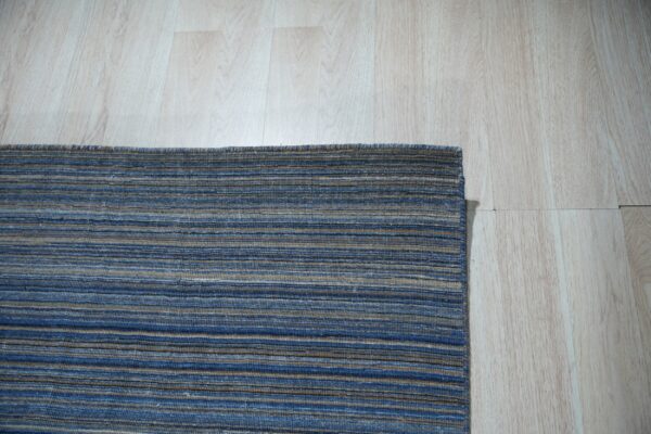 A corner view of a low-pile rug with blue, brown, and tan horizontal stripes on light wood flooring.