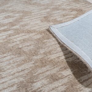 Beige and cream abstract rug showing its backing and bound edge on light wood flooring.