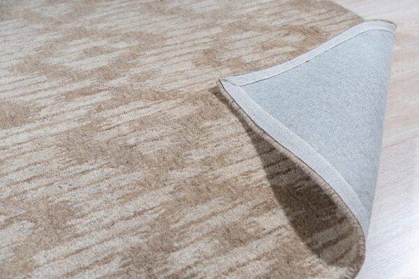 Beige and cream abstract rug showing its backing and bound edge on light wood flooring.