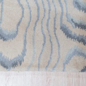 High-pile rug with abstract beige, blue, and gray wavy stripes on light plank flooring.