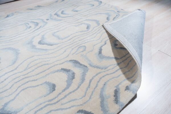 Cream and gray abstract rug rolled back on light wood flooring, showing the fabric backing.