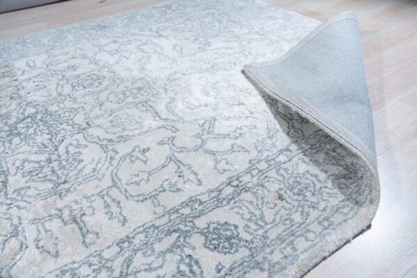 Cream rug with a faded blue ornate pattern lies on light wood flooring, revealing a gray fabric backing.