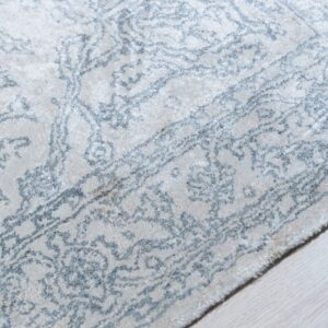 Textured ivory rug featuring a faded blue-gray ornate pattern shown against pale wood flooring.