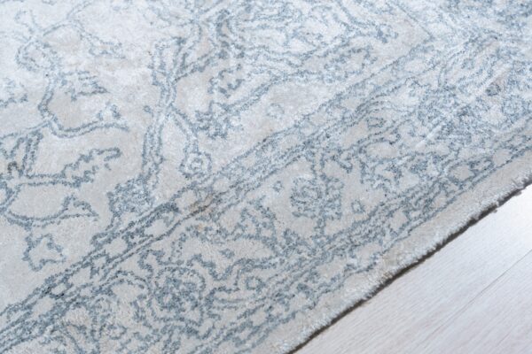 Textured ivory rug featuring a faded blue-gray ornate pattern shown against pale wood flooring.