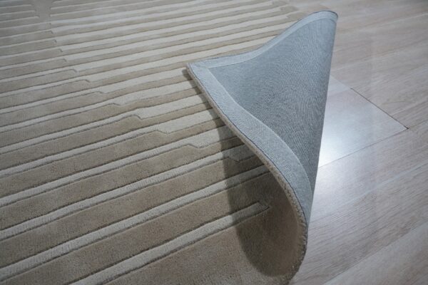 Beige striated rug partially lifted, showing a woven gray backing against pale wood flooring.